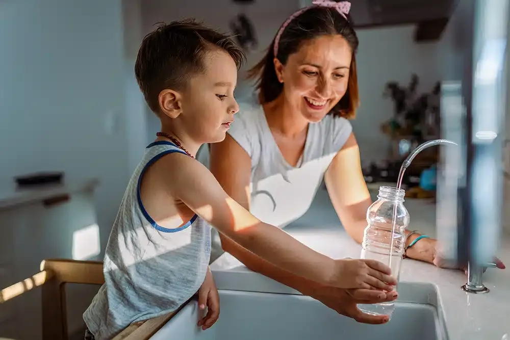 Mom helping son fill water bottle at kitchen sink in Hillsdale, NJ.